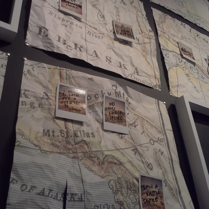 Wrinkled square maps tacked to a black wall, Polaroid photos with hand-written text scrawled on them are taped in various places on maps